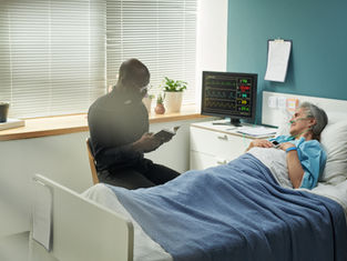 Elderly woman lies in hospital bed, connected to monitor. Clergyman reads beside her. Calm room with plants and blinds.