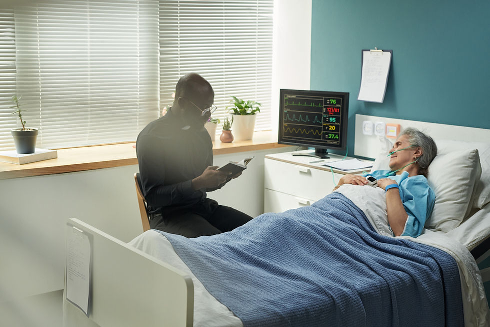 Elderly woman lies in hospital bed, connected to monitor. Clergyman reads beside her. Calm room with plants and blinds.