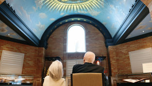 Two people sit in a room with a blue, decorative dome ceiling. They face a large arched window, surrounded by brick walls.