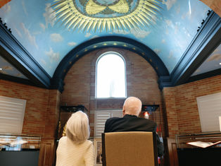 Two people sit in a room with a blue, decorative dome ceiling. They face a large arched window, surrounded by brick walls.