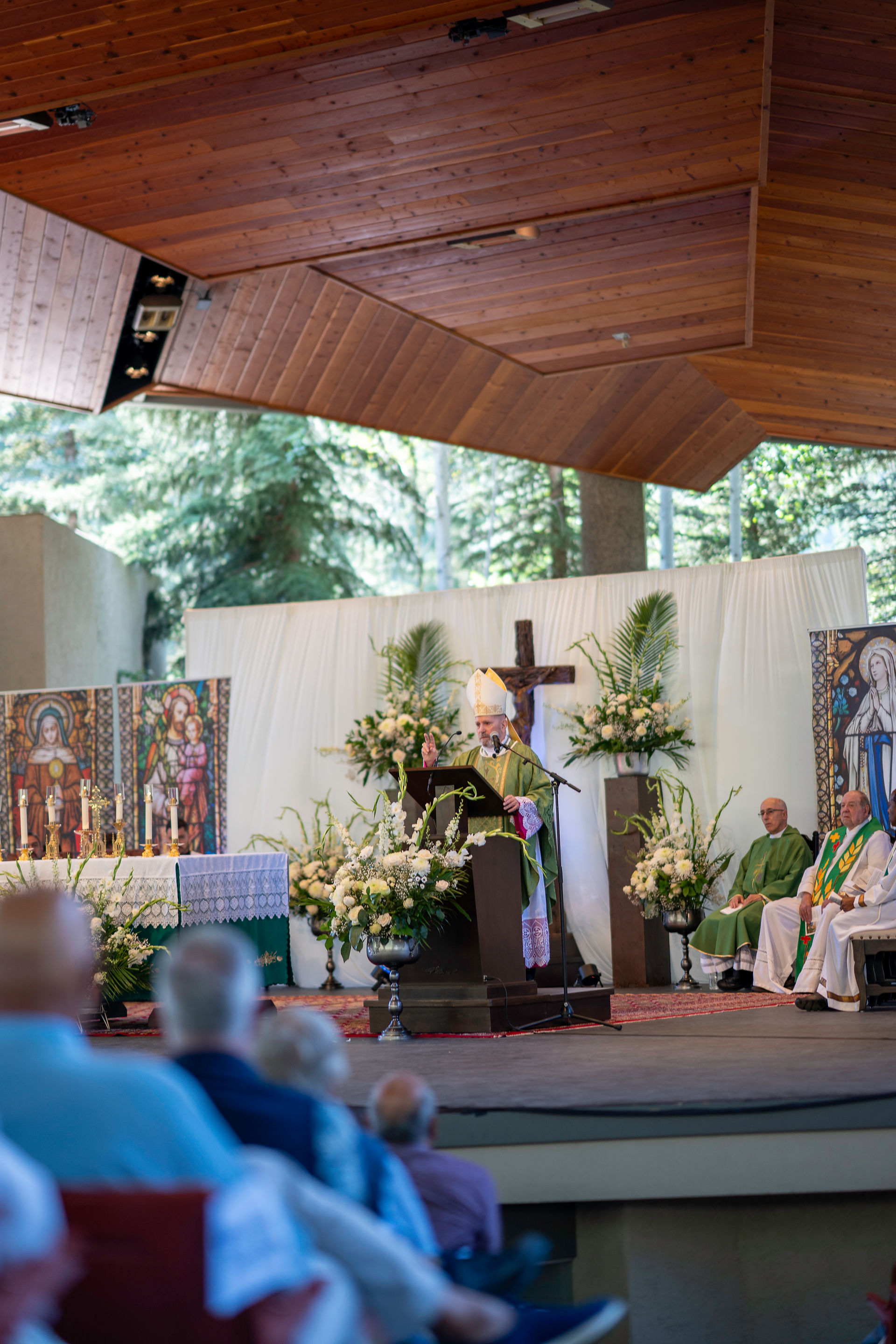 A bishop in green vestments speaks at a podium in a church with floral decor, stained glass art, and a cross. Audience members are seated.