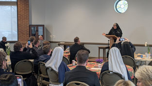 A nun speaks at a podium in a room with attendees seated at tables. Some guests take photos with phones. The mood is attentive.