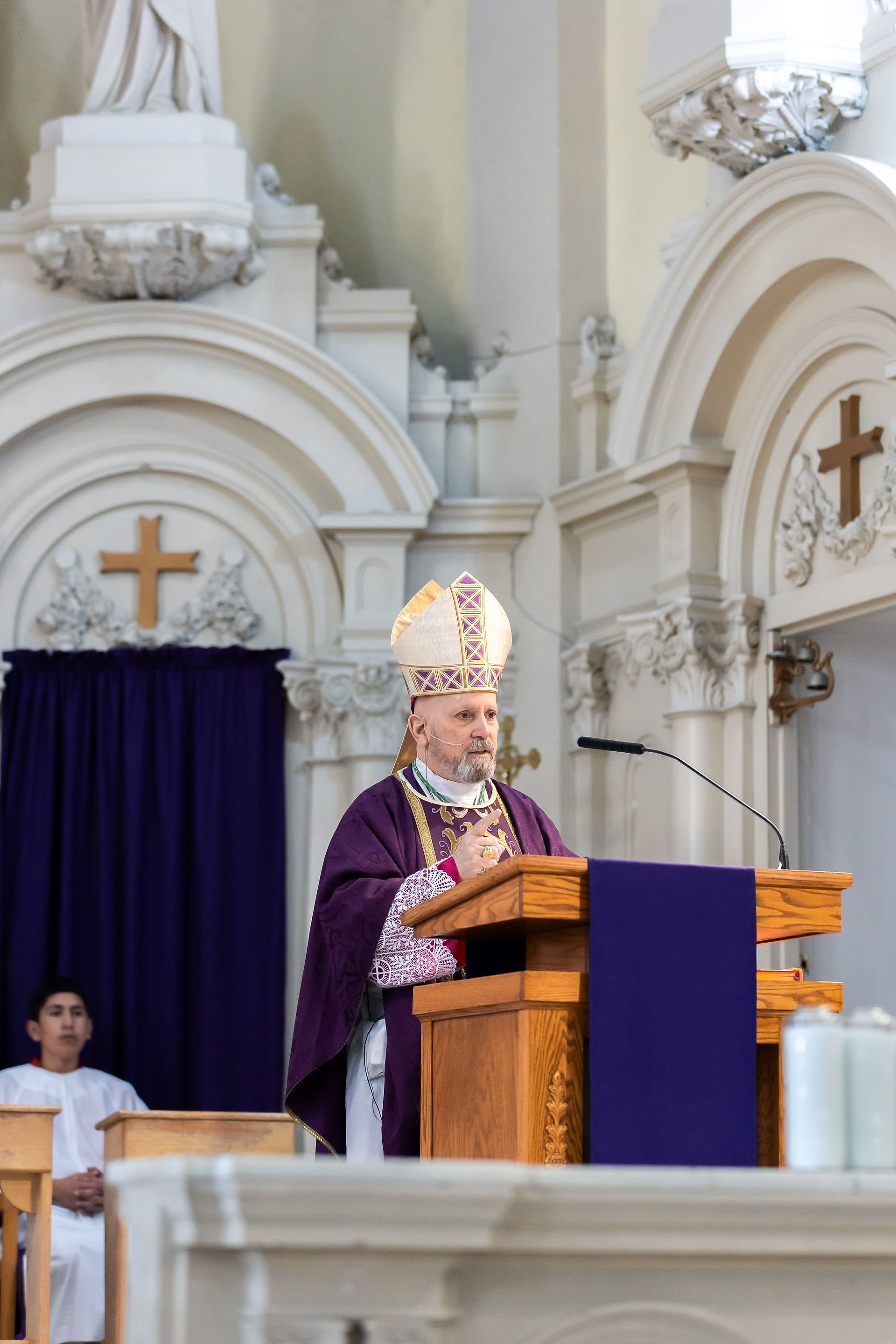 Bishop in purple and white attire at a church podium, speaking. Ornate background with crosses and a dark blue curtain. Peaceful setting.