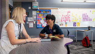 Teacher and student laugh while discussing a book at a table. Colorful sticky notes fill the classroom wall. Room 103 is visible.