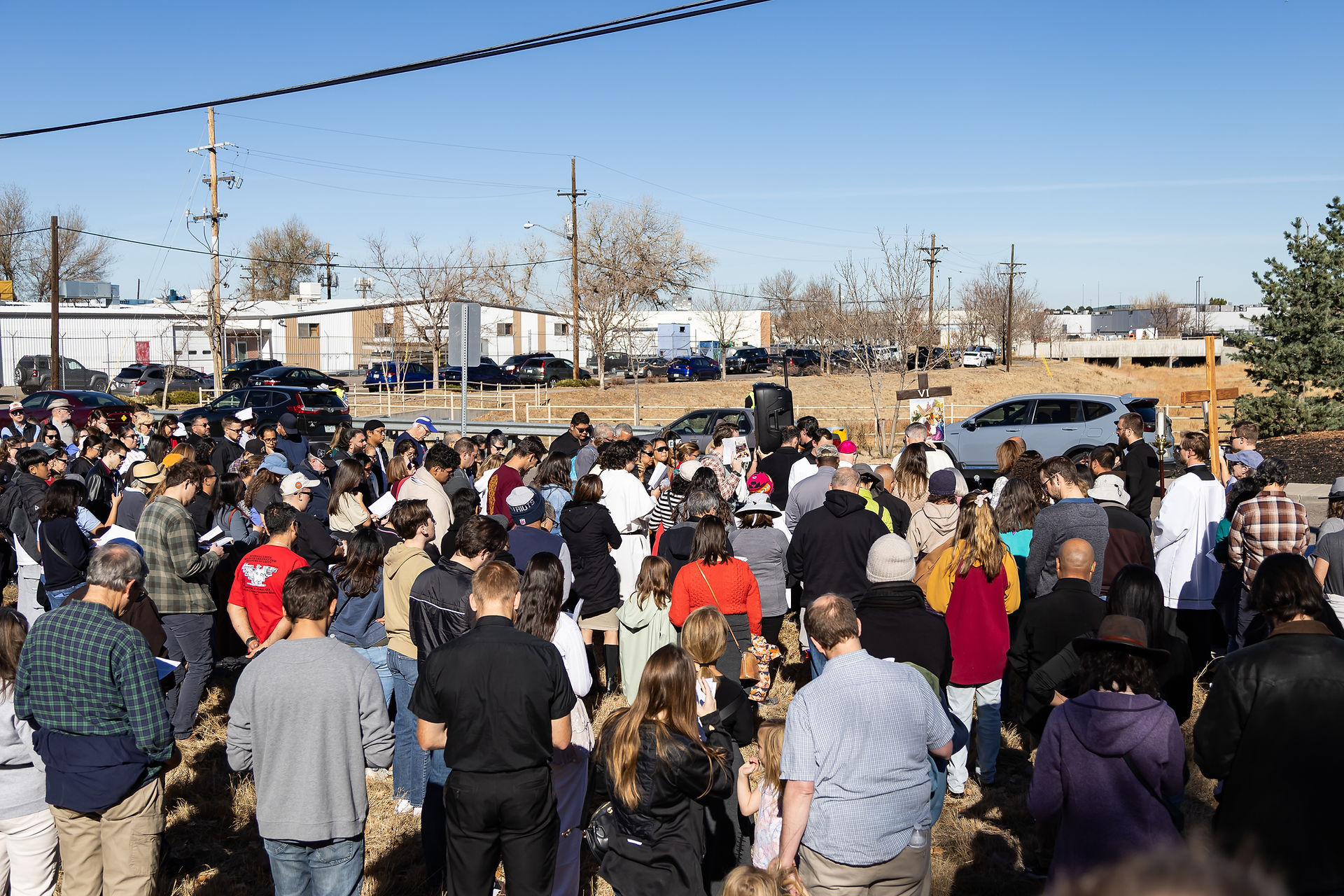 Crowd gathered outdoors for an event, some holding papers. Clear sky, trees, and buildings in the background. A cross marked "VI" visible.