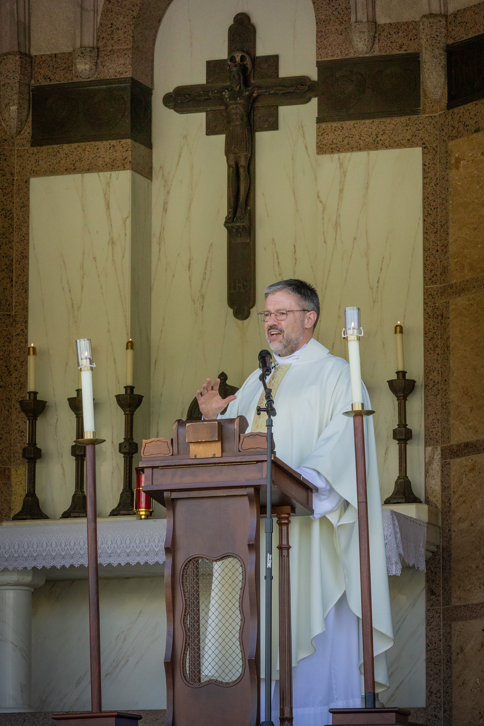 Priest in white robe speaks at a wooden lectern with candles in a church. A crucifix is visible on the wall behind him.