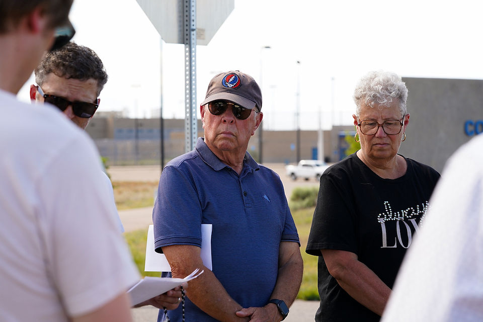 Older adults stand outdoors in a solemn group, one in a blue polo and cap, another in a black Always Love shirt, listening near a sign.