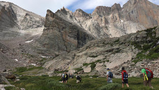 A group of hikers walks through an alpine meadow surrounded by dramatic rocky peaks and steep granite cliffs in a mountain wilderness.