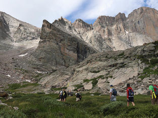 A group of hikers walks through an alpine meadow surrounded by dramatic rocky peaks and steep granite cliffs in a mountain wilderness.