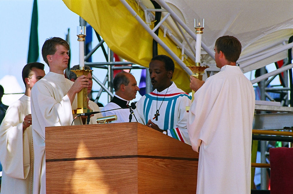 Deacon in white and colorful robes at a wooden podium hold candles during an outdoor ceremony. Bright flags hang in the background.