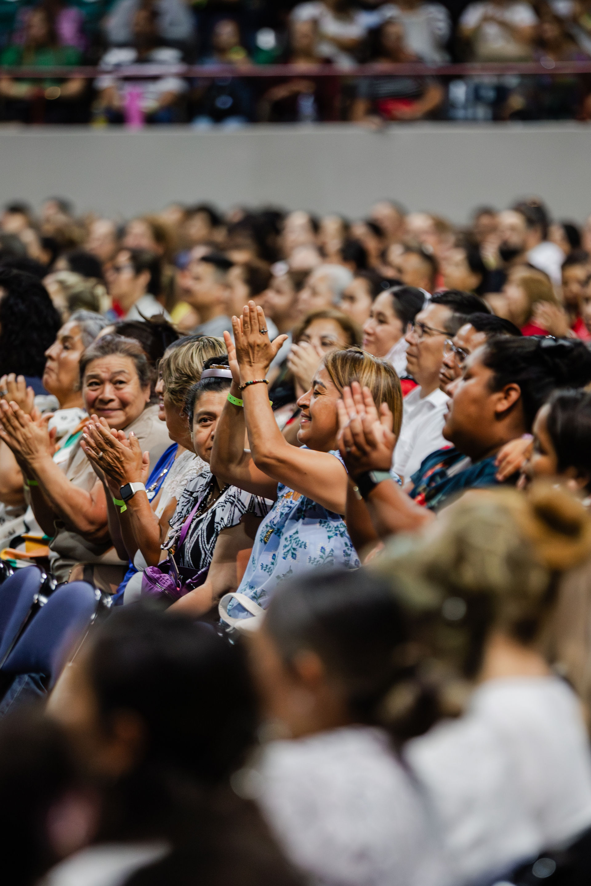 Audience clapping at an indoor event. People are smiling, showing excitement. Crowd fills rows of seats. Bright, lively atmosphere.