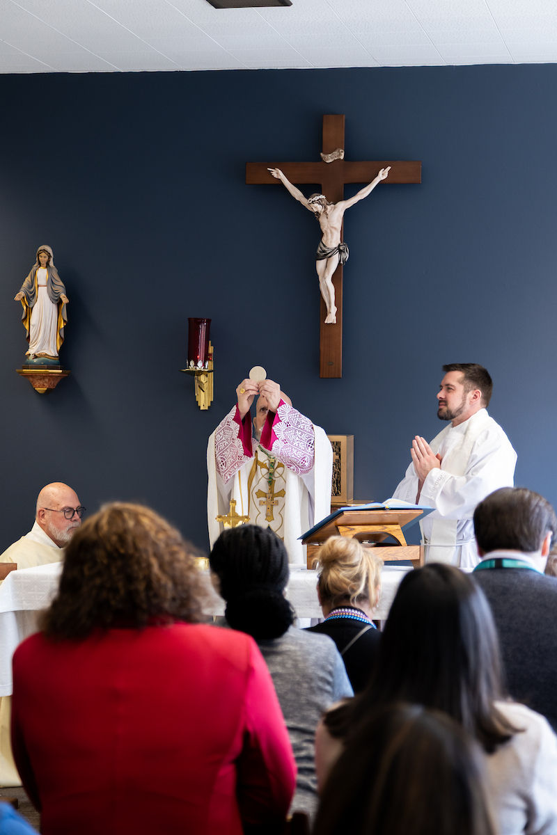 Priests conducting a mass, with one holding up the Eucharist. Blue wall with crucifix and statue. Congregation in foreground. Reverent atmosphere.