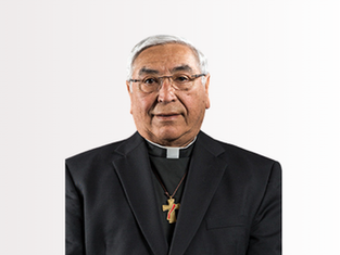 Man in a black clerical suit with a cross necklace, neutral expression, white background.