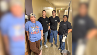 Four women stand in a hallway, smiling. One wears a blue "USA" shirt; others wear black shirts with "BD." A bucket and paper towels are nearby.