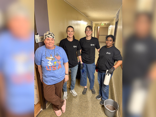 Four women stand in a hallway, smiling. One wears a blue "USA" shirt; others wear black shirts with "BD." A bucket and paper towels are nearby.
