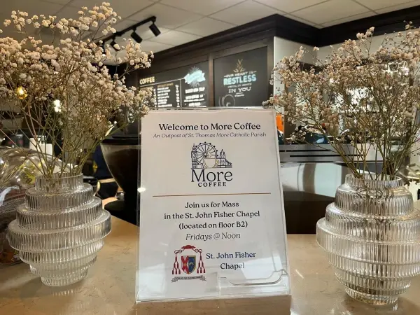 Coffee shop with two vases of white flowers. Sign reads: "Welcome to More Coffee...Join us for Mass...Fridays @ Noon. St. John Fisher Chapel."