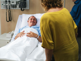 Elderly man smiling in a hospital bed, wearing a blue gown. A woman in a yellow shirt stands nearby. Medical equipment in the background.
