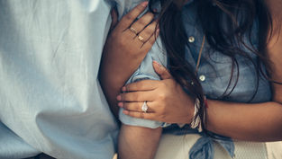 Close-up of two people in blue shirts with entwined arms, focusing on hands adorned with rings, conveying intimacy and connection.