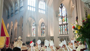 Priests in white robes sit in a cathedral with stained glass windows. Sunlight streams in, creating a serene atmosphere.