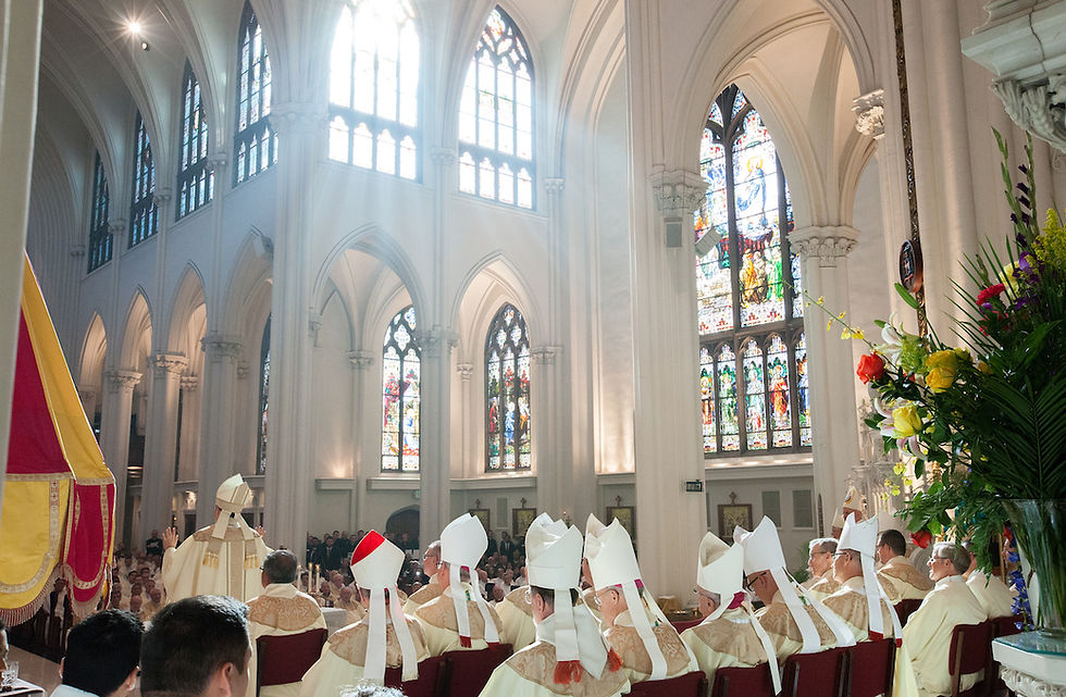 Priests in white robes sit in a cathedral with stained glass windows. Sunlight streams in, creating a serene atmosphere.