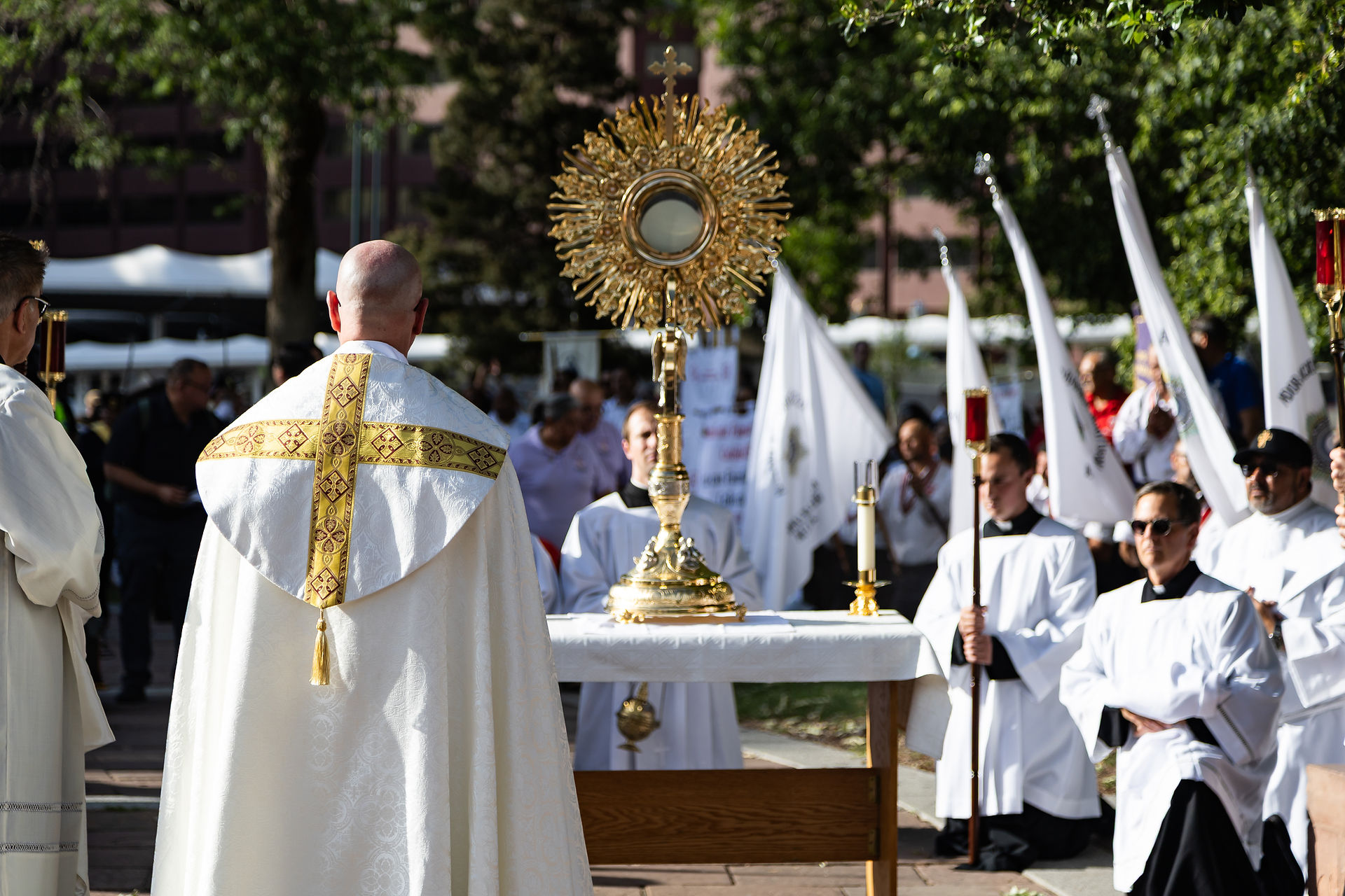 Priest in white and gold vestments leads an outdoor Catholic procession with a gold monstrance, altar servers, and flags.