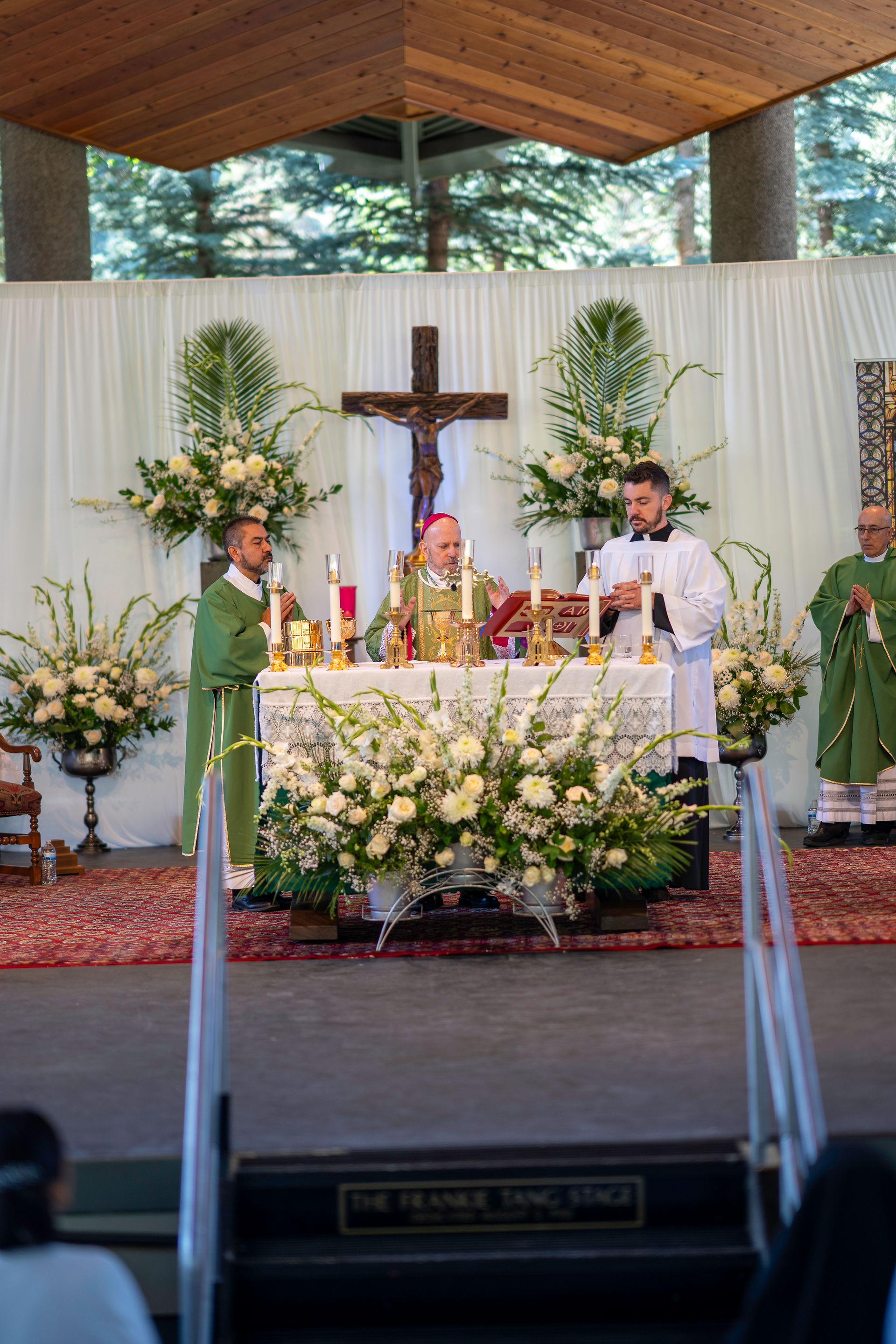 A religious ceremony with clergy at an altar, decorated with white flowers. A cross and greenery are in the background. Mood is solemn.
