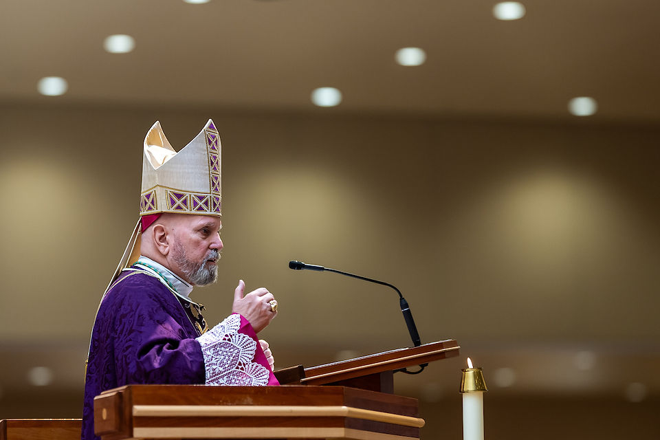 A bishop in purple robes and mitre speaks at a lectern with a microphone. Background is softly lit with circular lights.