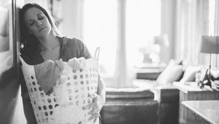 Woman holding a laundry basket leans tiredly against a wall in a softly lit room with a cozy sofa and cushions in the background. Monochrome.