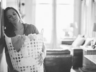 Woman holding a laundry basket leans tiredly against a wall in a softly lit room with a cozy sofa and cushions in the background. Monochrome.
