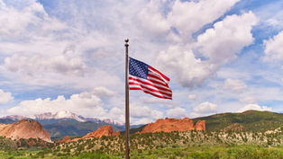 American flag on a pole above red rock mountains and green hills under a bright, cloudy blue sky.