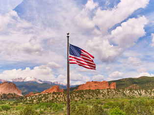 American flag on a pole above red rock mountains and green hills under a bright, cloudy blue sky.