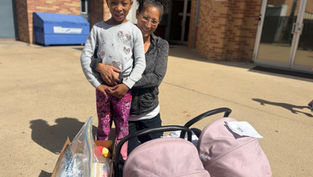 Woman and child smiling outside a brick building. They stand beside a box and pink stroller. Sunny day with shadows on the ground.
