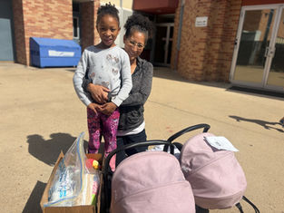 Woman and child smiling outside a brick building. They stand beside a box and pink stroller. Sunny day with shadows on the ground.