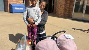 Woman and child smiling outside a brick building. They stand beside a box and pink stroller. Sunny day with shadows on the ground.