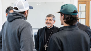 A smiling clergyman with a large cross necklace converses with three casually dressed men in caps inside a bright room.
