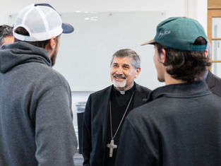 A smiling clergyman with a large cross necklace converses with three casually dressed men in caps inside a bright room.