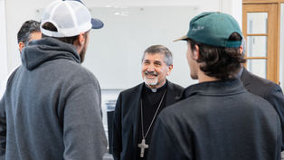 A smiling clergyman with a large cross necklace converses with three casually dressed men in caps inside a bright room.