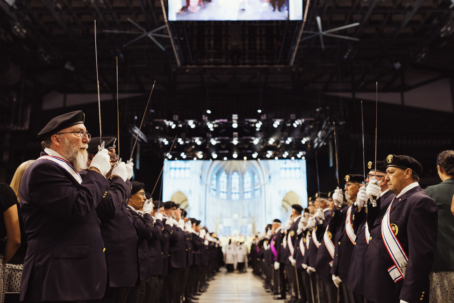 Uniformed men with berets hold swords upwards in an indoor ceremony with a grand, illuminated backdrop. Formal and solemn atmosphere.