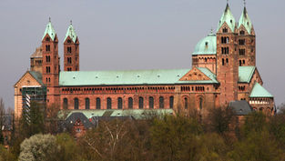 Large Romanesque cathedral with twin towers and green copper roofs above trees under a hazy sky