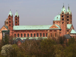 Large Romanesque cathedral with twin towers and green copper roofs above trees under a hazy sky