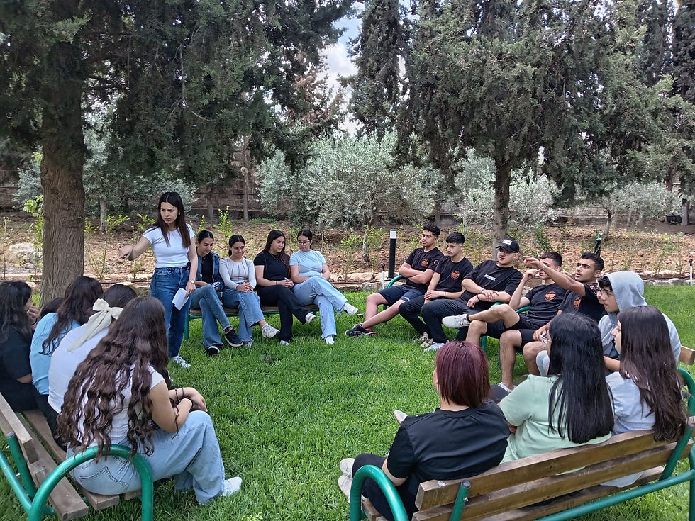 A group of people sit on benches in a park, engaged in a discussion. Tall trees and greenery create a relaxed outdoor setting.