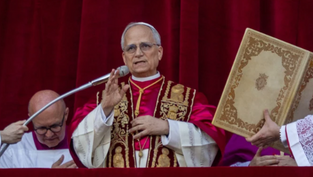 A man in ornate red and gold robes speaks into a microphone on a balcony, holding a gesture. Another holds a large book. Red curtain backdrop.