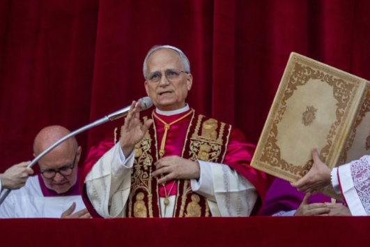 A man in ornate red and gold robes speaks into a microphone on a balcony, holding a gesture. Another holds a large book. Red curtain backdrop.