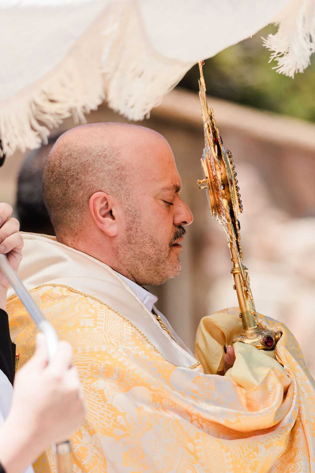 Bald man in ornate gold-and-cream robes holds a gilded religious icon, eyes closed, under a white canopy at a solemn ceremony.