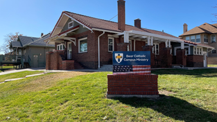Brick building with red-tiled roof and white trim labeled 'Bear Catholic Campus Ministry'; a blue sign with shield emblem and American flag design stands on the grassy lawn under a clear blue sky.