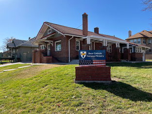 Brick building with red-tiled roof and white trim labeled 'Bear Catholic Campus Ministry'; a blue sign with shield emblem and American flag design stands on the grassy lawn under a clear blue sky.