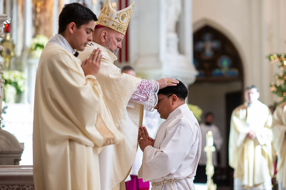 A bishop in ornate attire places his hand on a kneeling man's head in a church, surrounded by other clergy. The setting is solemn and reverent.