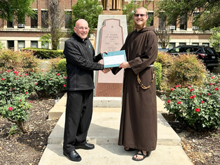 Two smiling men exchange a large check outdoors by a statue, surrounded by bushes and red flowers, with a building in the background.