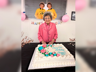 Elderly woman in pink stands smiling behind a birthday cake with floral decoration and "Happy 101st Birthday Peggy!" banner and balloons behind.