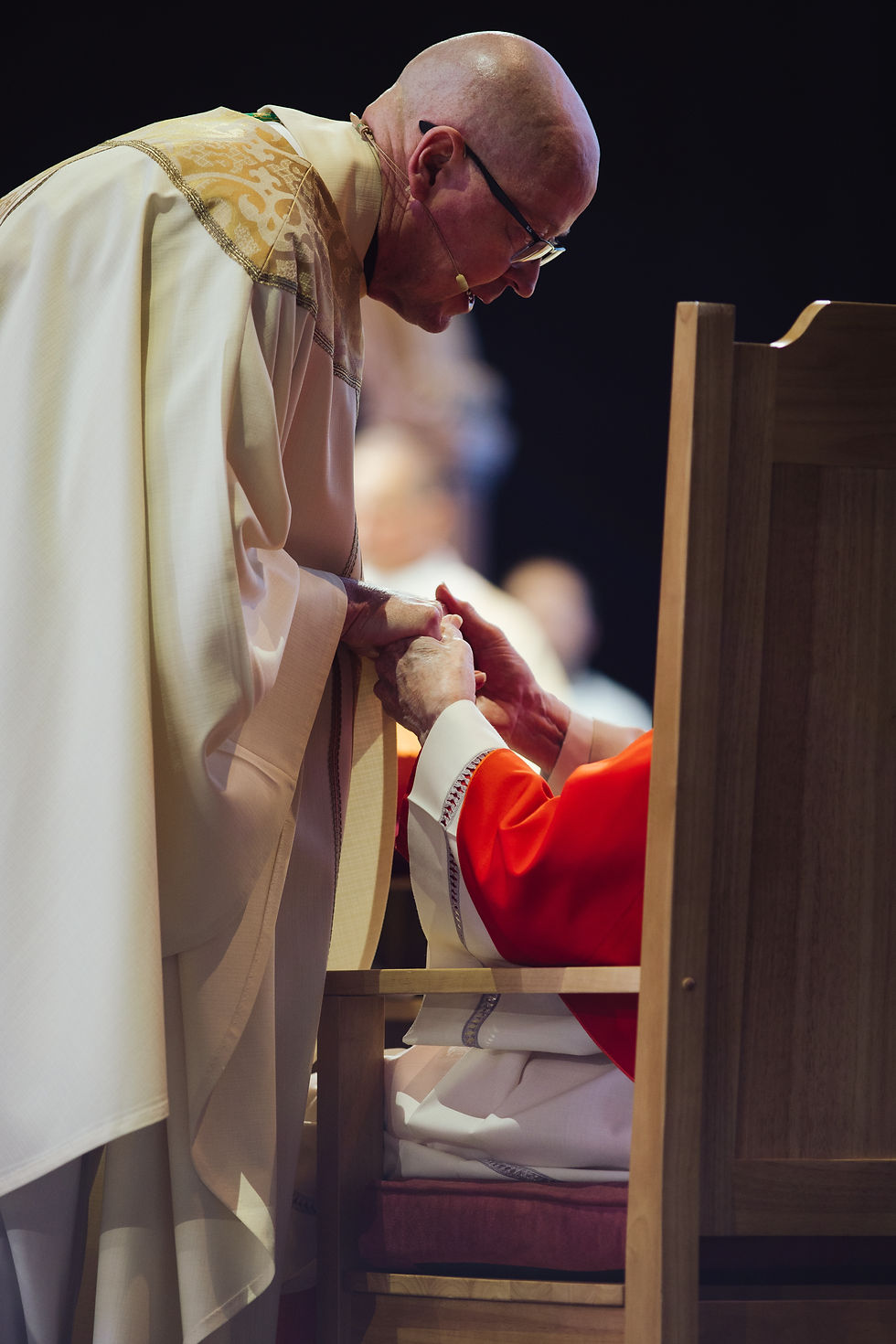 Priest in white robe holds the hands of seated person in red robe, conveying a solemn, respectful mood on a wooden chair background.